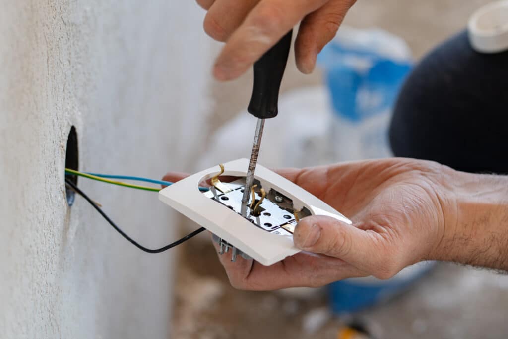 Close up on hands of caucasian man electrician holding screwdriver working on the plug electric on residential electric system installing white AC power socket on gray wall at home repair close up