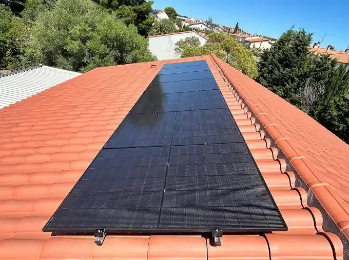 Panneaux solaires noirs sur un toit en tuiles orange, quartier résidentiel arboré sous un ciel bleu.