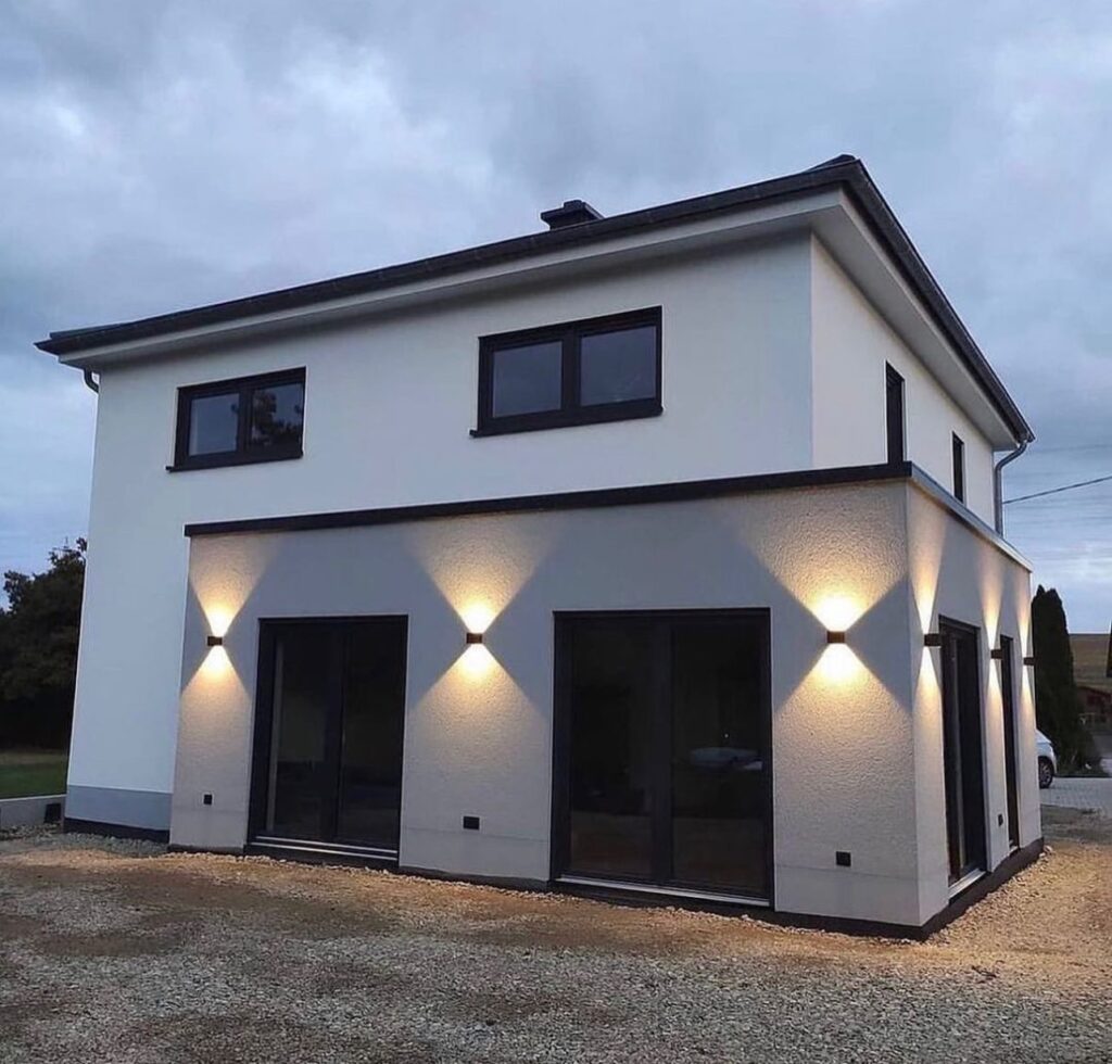 Maison Moderne : Design Extérieur et Éclairage de Façade Maison moderne bicolore avec éclairage mural design et baies vitrées sombres sur gravier au crépuscule.